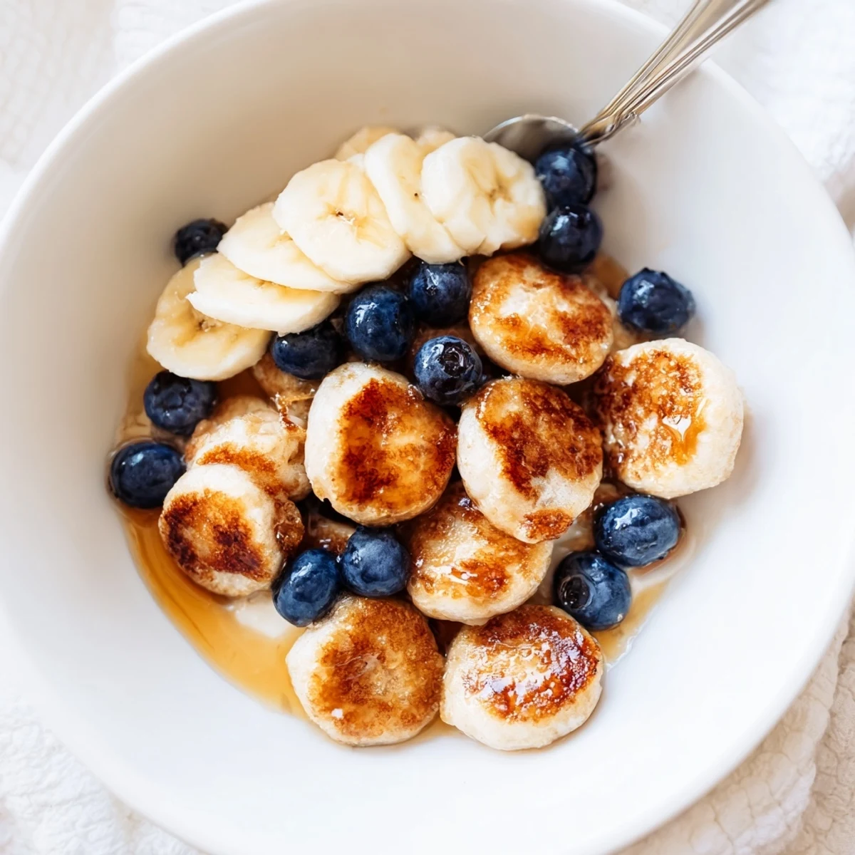 Fluffy mini pancake cereal topped with fruit, drizzled with maple syrup for sweetness.